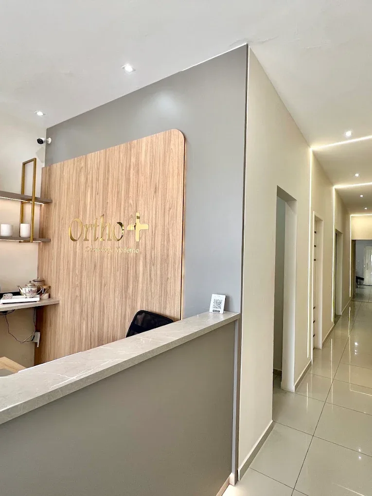 Recepção da Orthomais: painel de madeira com a logomarca em dourado, bancada de mármore claro e corredor de atendimento iluminado ao fundo.